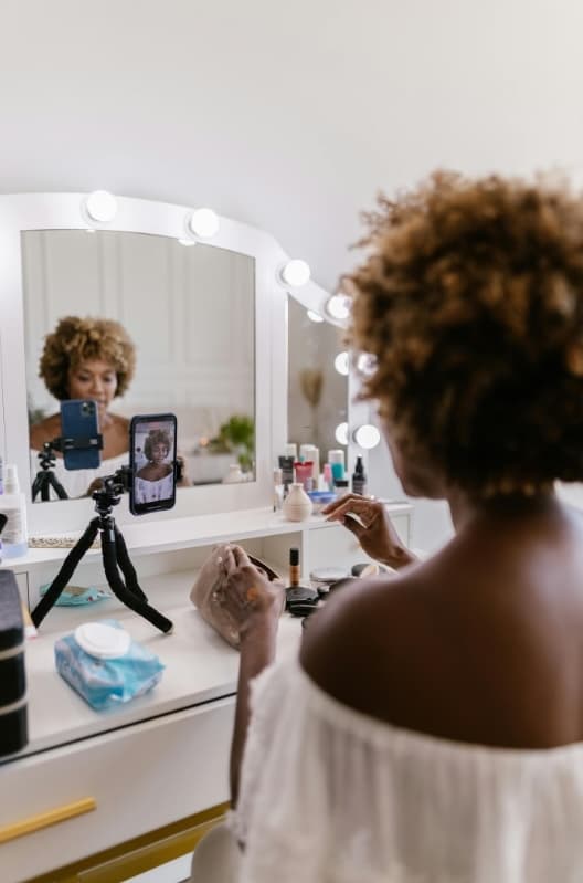 Lash tech recording an Instagram Reel in front of a lit vanity mirror with her phone on a tripod