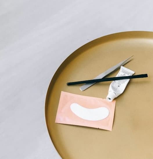 Lash extension supplies laid out on a gold tray during a quiet slow-season week: isolation tweezers, a lash tool, adhesive, and under-eye gel patches