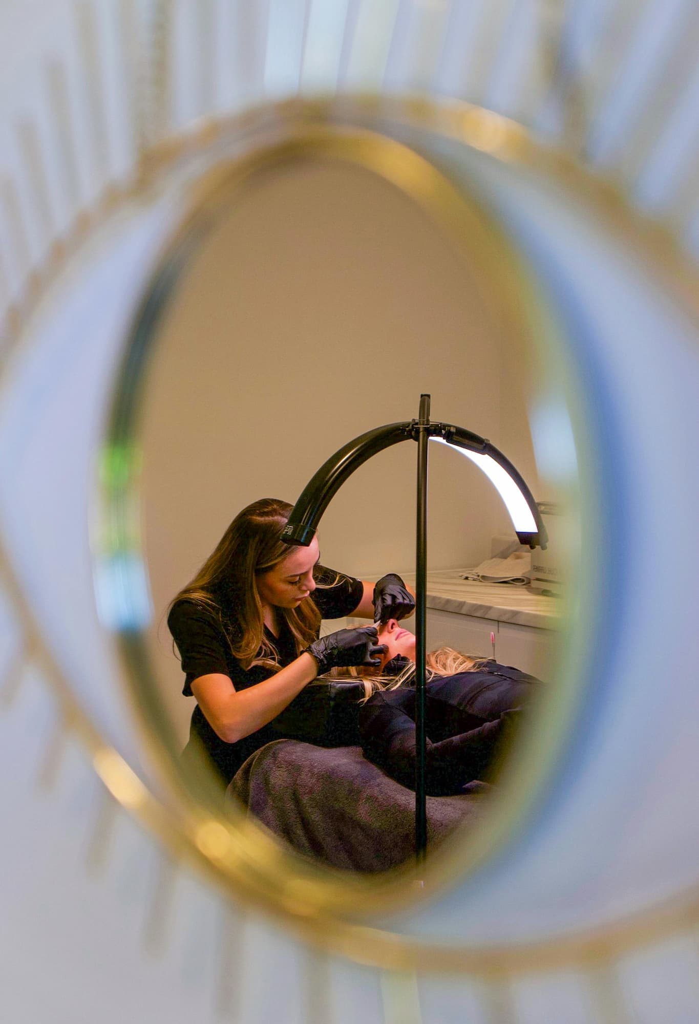 Lash tech applying lash extensions to a client seen through a mirror reflection